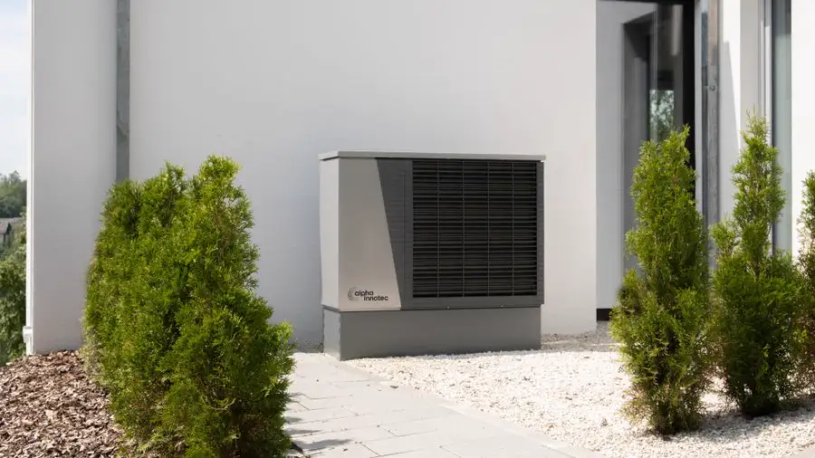 Large gray outdoor air conditioner or heat pump unit beside a white house wall with small evergreen shrubs