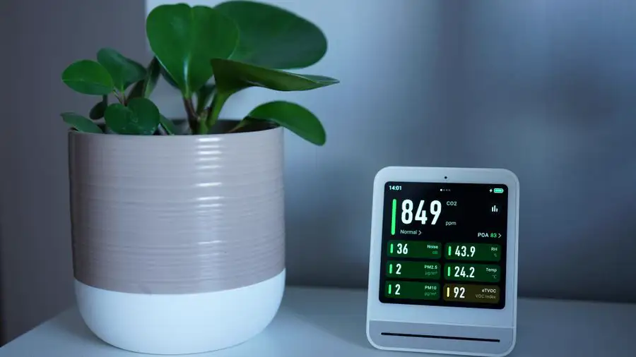 Small indoor air monitor showing humidity and air readings beside a potted plant