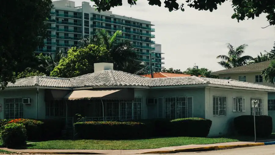 Low pastel house with a tile roof and a window air conditioner, palms and a tall building behind