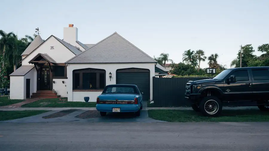 White house with a gray roof and garage, cars parked in front, palm trees behind