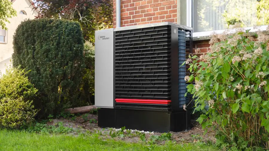 Gray outdoor heat pump unit beside a house with shrubs and a brick wall