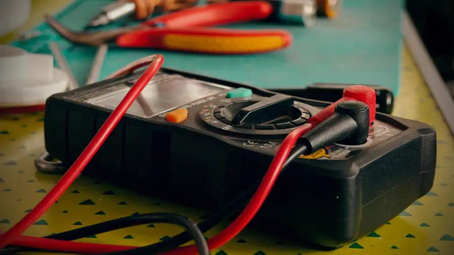 Multimeter with red and black test leads and pliers on a work table, the kind of tools used to test an electrical part