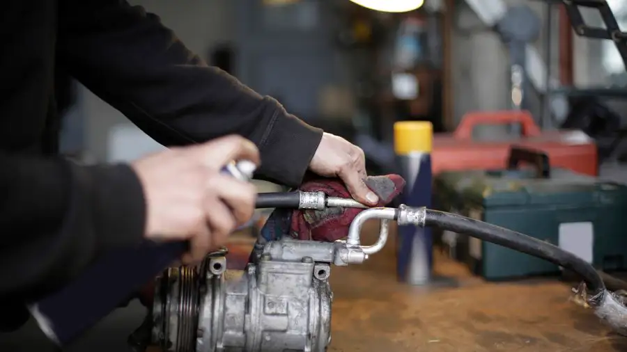 Hands connecting a hose to an air conditioning compressor on a workbench