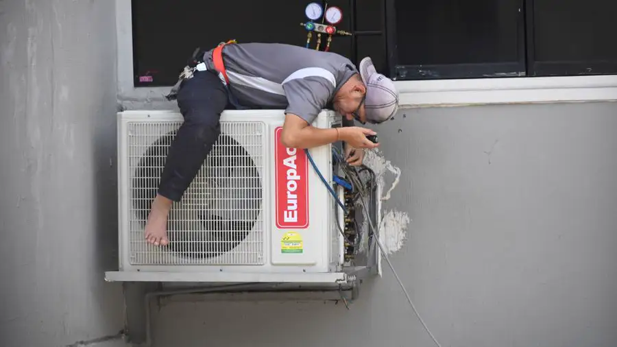 Technician leaning out to repair an outdoor air conditioner mounted on a building wall