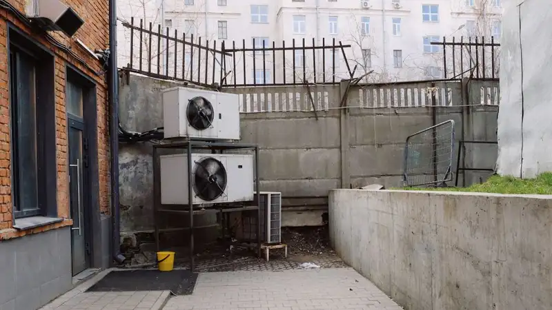 Air conditioning system located outside concrete shabby fence with metal barrier near industrial building from bricks and glass door