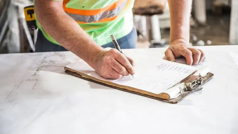 A construction worker writes notes on a clipboard at a construction site, emphasizing planning and precision