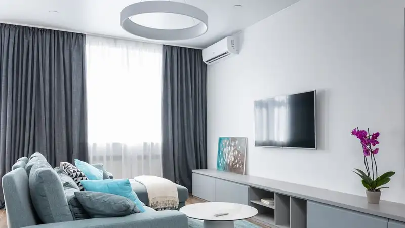 Living room with a white wall-mounted split air conditioner high on the wall beside a wall-mounted TV
